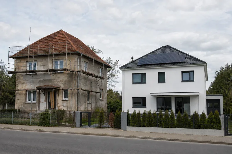 Sanierungsbedürftiger Altbau mit Gerüst neben modernem weißem Neubau mit Photovoltaik (KI-generiertes Symbolbild)